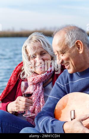 Glückliches Seniorenpaar, das die Zeit zusammen genießt, Gitarre spielt und Wein am See trinkt, umhüllt in einer roten Decke. Stockfoto