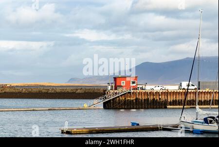 Pier mit Sicherheitskabine und Leiter, während die Fahrzeuge auf dem Damm geparkt sind und das Boot mit einem Segelstock, der auf dem plätschernden Meerwasser gegen den Berg schwimmt Stockfoto