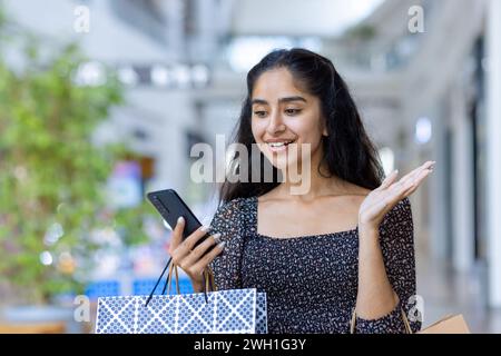 Fröhliche indische Käuferin, die ein Handy benutzt, während sie bunte Taschen in einem modernen Einkaufszentrum hält. Stockfoto