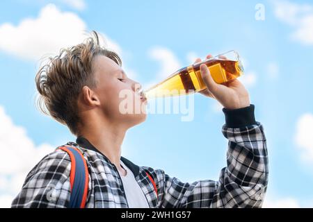 Fröhlicher Junge, der Limonade aus einer Glasflasche vor einer wunderschönen Himmelskulisse trinkt. Er trägt ein kariertes Hemd und einen Rucksack. Lebendige Farben Stockfoto