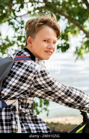 Porträt eines lächelnden Jungen mit blonden Haaren im karierten Hemd mit Rucksack, der auf dem Fahrrad sitzt, auf verschwommenen Bäumen und reflektierendem Teichhintergrund am sonnigen Tag Stockfoto