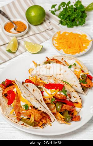 Gebackene Tex-mex-Hühnchen-Fajitas mit gemischtem süßem Pfeffer, Zwiebeln, Sauerrahm, geriebenem Käse und weißen Maistortillas auf Teller auf weißem Holztisch mit Stockfoto