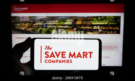 Person, die ein Mobiltelefon mit dem Logo des US-Supermarktunternehmens The Save Mart Companies vor der Business-Webseite hält. Schwerpunkt auf dem Display des Telefons. Stockfoto