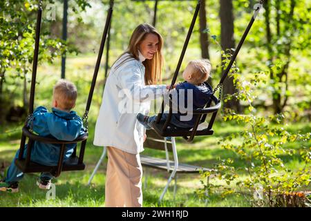 Die Mutter mit zwei Kindern macht Spaß auf dem Spielplatz. Die Mutter schwingt ihre Kinder auf den Schaukeln. Stockfoto