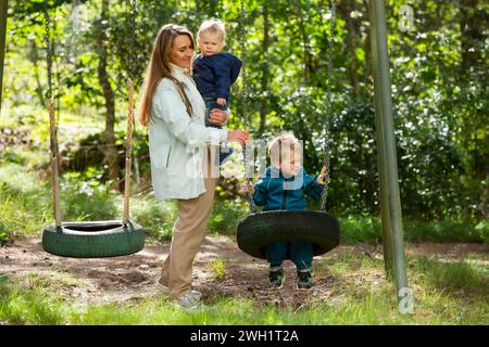 Die Mutter mit zwei Kindern macht Spaß auf dem Spielplatz. Die Mutter schwingt ihre Kinder auf den Schaukeln. Stockfoto