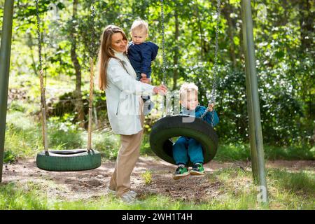 Die Mutter mit zwei Kindern macht Spaß auf dem Spielplatz. Die Mutter schwingt ihre Kinder auf den Schaukeln. Stockfoto