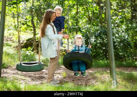 Die Mutter mit zwei Kindern macht Spaß auf dem Spielplatz. Die Mutter schwingt ihre Kinder auf den Schaukeln. Stockfoto