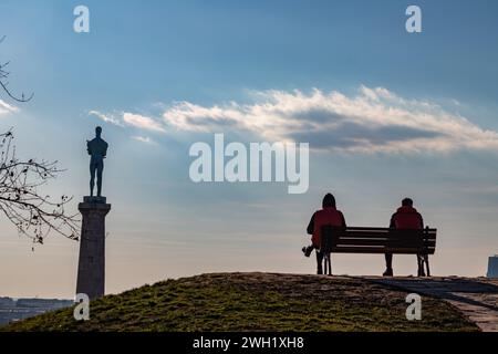 Zwei Freunde draußen an einem schönen sonnigen Tag, sitzen auf der Bank der Kalemegdan-Festung in Belgrad, mit wunderschönem Blick auf die Siegesstatue (Pobednik) Stockfoto