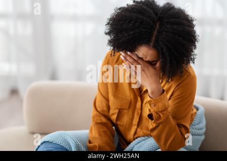 Eine junge Frau in einem senfgelben Hemd sitzt auf einem beigen Sofa und bedeckt ihr Gesicht Stockfoto