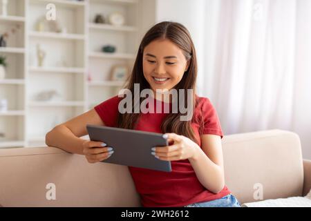 Asiatische Frau, die sich mit einem digitalen Tablet entspannt, während sie bequem auf einer modernen Couch sitzt Stockfoto