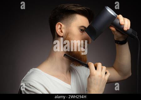 Porträt eines langbärtigen Mannes mit Haarfön und Kamm zu Hause, dunkler Hintergrund, Nahaufnahme Stockfoto