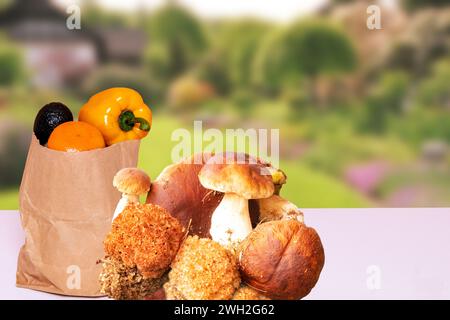 Verschiedene Arten von essbaren Waldpilzen wie Stachelschweine, Sonnenschirme und eine braune Papiertüte voller frischem, gesundem Gemüse auf dem Tisch über einer verschwommenen Natu Stockfoto