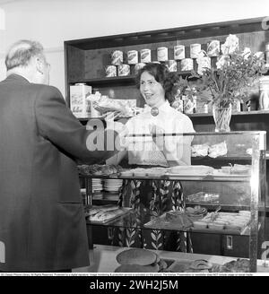 Die Konditorei in den 1950er Jahren Eine Konditorei in Eskilstuna, in der ein männlicher Kunde steht und an der gut sortierten Theke mit Gebäck, Brötchen und Kuchen kauft. Eine junge Frau arbeitet in der Konditorei und sieht glücklich aus, als sie dem männlichen Kunden von der Auswahl an Gebäck erzählt. 3. Juni 1954. Conard Ref. 2687 Stockfoto