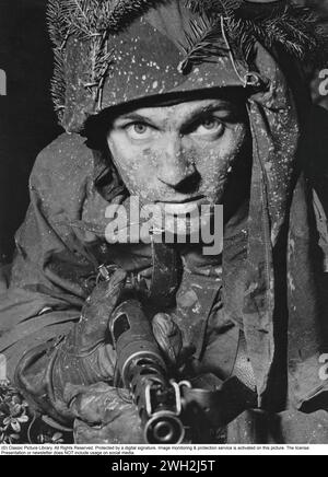 Schwedischer Soldat in den 1950er Jahren Ein junger Mann hat sich während einer Militäraktion gut mit Tannenzweigen im Helm getarnt. Er hat eine geladene Maschinenpistole in der Hand und ist bereit für den Kampf. 1945 führte Schweden das 9-mm-Parabellum Carl Gustaf m/45 ein, dessen Design viele Konstruktionselemente früherer Maschinenpistolen aufnahm und diese verbessert. Er verfügt über einen rohrförmigen gestanzten Stahlbehälter mit seitlichem Faltschaft. Die m/45 wurde weit verbreitet exportiert und besonders beliebt bei CIA-Agenten und US-Sondereinheiten während des Vietnamkriegs. In den USA war es als Schwedisch-K bekannt Stockfoto