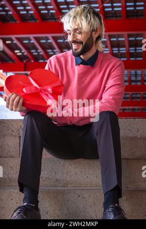 Ein junger, lächelnder Mann mit vertikalem Schuss hält eine herzförmige Geschenkbox. Liebhaber Stockfoto