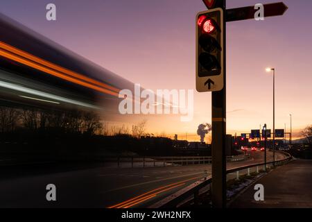 Leichte Busspuren, die an einer roten Ampel auf einer Straße vorbeifahren. Magentafarbenes Nachglühen im Hintergrund, hinter Ampeln und Richtungsschildern. Langzeitbelichtung. Stockfoto