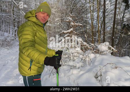 Der Mann hat Daunenjacke angezogen, im Wald im Winter mit Trekkingstöcken und Schneeschuhen, sonniger Morgen nach Schneefall. Stockfoto