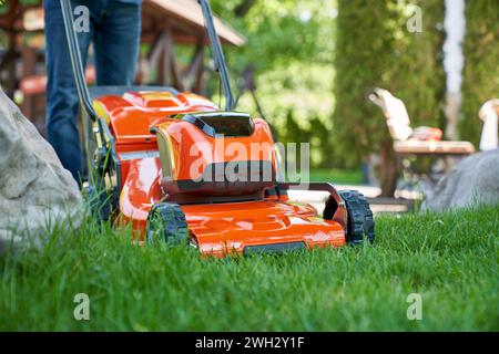 Bildausschnitt eines nicht erkennbaren Gärtners in Jeans mit Rasenmäher im Hinterhof. Nahaufnahme des orangefarbenen Rasentrimmers, der frisches grünes Gras schneidet, mit verschwommenem Mann im Hintergrund. Konzept der Gartenausrüstung. Stockfoto