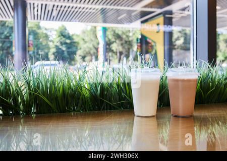 Milchshake zum Mitnehmen, Coffee Shop bei Tankstelle. Zwei durchsichtige Plastikbecher mit Deckel gefüllt Schokoladen-Vanille-Milchshakes auf dem Tisch, grünes Gras. Bla Stockfoto