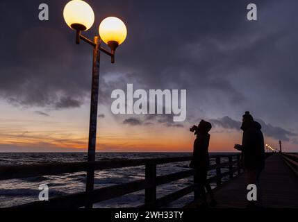 Wustrow, Deutschland. Februar 2024. Eine Frau fotografiert den Sonnenuntergang über der Ostsee auf dem Pier. Mit kalten Temperaturen von rund drei Grad und Sturmwind zeigt das Wetter in Norddeutschland seine winterliche Seite. Quelle: Jens Büttner/dpa/Alamy Live News Stockfoto