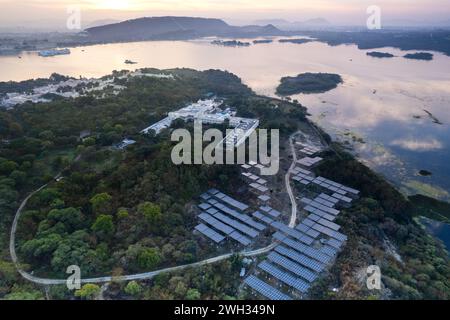 Luftdrohne mit Solarpaneelen, die in der Mitte von Bäumen am Seeufer platziert wurden, reflektieren die Farben des Sonnenaufgangs und aravalli-Hügel, die grene ökologische Re zeigen Stockfoto