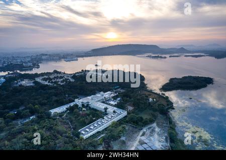 Drohnenaufnahme mit einem großen Resort-Hotel auf einem baumbedeckten Ufer mit Blick auf den See pichola fateh sagar mit Sonnenaufgang über den aravalli-Hügeln udaipur, Jodhpur Stockfoto