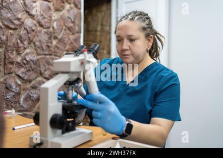 Eine fokussierte Labortechnikerin in blauen Peelings und Handschuhen analysiert die Probe sorgfältig mit dem Labormikroskop. Stockfoto