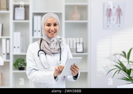 Schöne arabische medizinische Frau, die im Arbeitsbereich mit digitalem Tablet in den Händen steht. Selbstbewusste Frau mit Stethoskop aussehender Kamera mit Lächeln und Einsatz von Gadget für die Kommunikation mit Patienten. Stockfoto