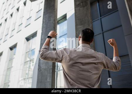 Rückansicht eines brünetten Mannes im Hemd, der ein Powerschild mit geballten Fäusten macht, während er neben dem Bürogebäude steht. Ein nicht erkennbarer Kerl fühlt sich stark und mächtig, nachdem er für einen Traumjob eingestellt wurde. Stockfoto