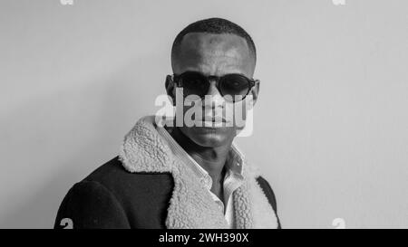 Ein Mann steht mit einer Sonnenbrille und einer blauen Jacke und blickt auf die Kamera über ein helles, einfarbiges, hochauflösendes Hintergrundbild Stockfoto