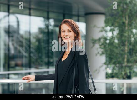 Porträt einer fröhlichen Frau mittleren Alters in schwarzer formeller Kleidung. Geschäftsfrau mit Ingwerhaaren am Geländer draußen. Stockfoto