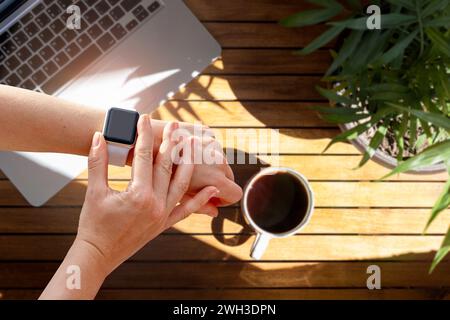 Intelligente Uhr auf der Hand einer Frau über einem Holztisch mit Laptop und einer Tasse Kaffee. Stockfoto