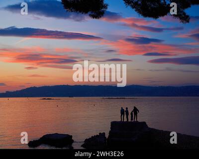 Bewölkter Sonnenuntergang über der Bucht von Cannes, Frankreich, mit Menschen auf den Felsen, die vor dem ruhigen rosa Meer stehen. Stockfoto