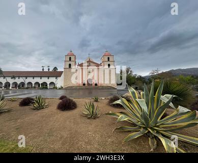 Santa Barbara, CA, USA - 18. Dezember 2023: Lange Sicht, grüne, gelbe Kakteen vor der Old Mission Kirche unter grauer Wolkenlandschaft. Berge und Bäume auf Stockfoto