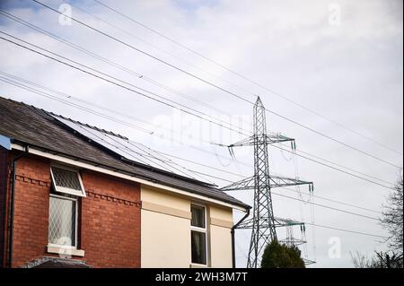 Haus mit Sonnenkollektoren auf dem Dach unter nationalen Stromleitungen und Pylon mit zwei Formen der Stromerzeugung Stockfoto