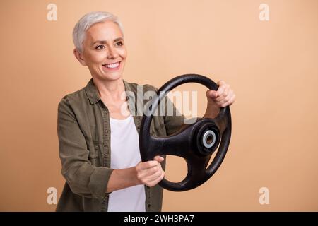 Porträt einer fröhlichen blonden Kurzhaarfrau im Simulatorfahrer, der das Lenkrad-Spielgerät isoliert auf beigefarbenem Hintergrund hält Stockfoto