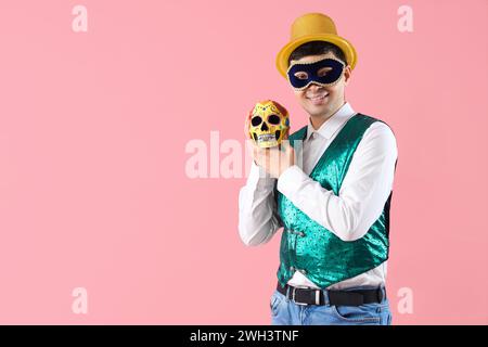 Glücklicher junger Mann in Kostüm und Karnevalsmaske, der gemalten menschlichen Schädel auf rosa Hintergrund hält Stockfoto