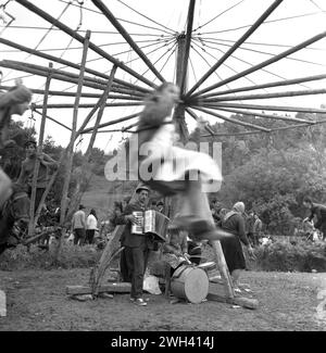 Vrancea County, Rumänien, ca. 1977. Einheimische genießen eine Fahrt auf dem Landmarkt in den Kettenschaukeln, die von einem Pferd gedreht werden, mit einem kleinen Musikensemble, das für Unterhaltung bezahlt wird. Stockfoto