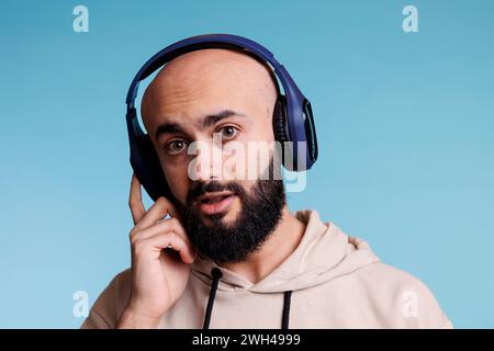 Arabischer Mann, der Musik in kabellosen Kopfhörern genießt und in die Kamera schaut. Junge Person, die Ohrhörer berührt, während sie Songs Playlist Studio-Porträt auf blauem Hintergrund anhört Stockfoto