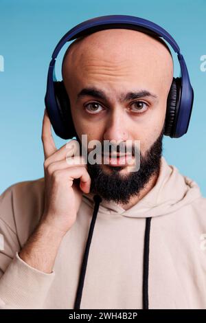 Junger arabischer Mann, der Musik im Porträt kabelloser Kopfhörer hört. Glatzbärtige Person, die Musik-Playlist in Ohrhörern genießt, während sie mit neutralem Ausdruck in die Kamera schaut Stockfoto