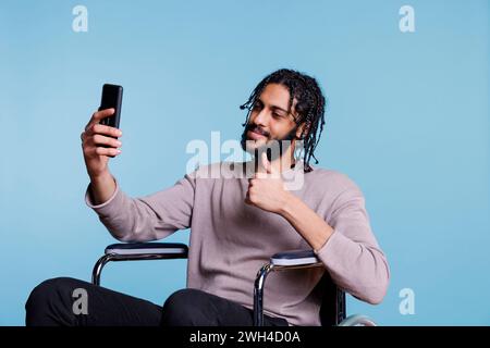 Junger arabischer Mann, der in Videogesprächen auf dem Smartphone spricht und im Rollstuhl den Daumen nach oben zeigt. Person mit körperlicher Behinderung, die Online-Kommunikation auf dem Mobiltelefon hat Stockfoto
