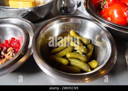 Eine Schüssel aus Edelstahl voller knuspriger Gurken zum Servieren, umgeben von anderen Zutaten in einer belebten Küche. Stockfoto