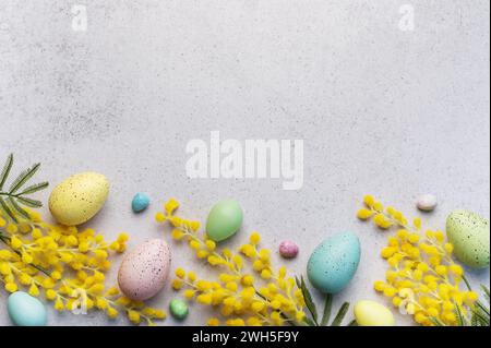 Dieses Bild zeigt ein festliches Osterarrangement aus pastellfarbenen Eiern und hellgelben Mimosa-Blüten auf einer strukturierten hellgrauen Oberfläche. Stockfoto