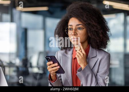 Konzentrierte Unternehmerin mit lockigem Haar und Brille, die in einem Firmenumfeld ein Telefon hält. Stockfoto