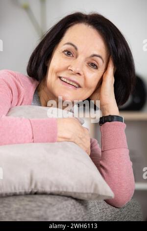 Reizende Seniorin, die auf einem Sofa sitzt und ein Kissen kuschelt Stockfoto