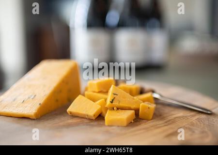 Eine Nahaufnahme von Cheddar-Käse, geschnitten auf einem Olivenholzbrett mit einer Weinflasche im Hintergrund Stockfoto