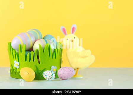 Frohe Ostern. Farbige ostereier in einem grünen Korb neben einem Küken mit Kaninchenohren auf gelbem Hintergrund Stockfoto