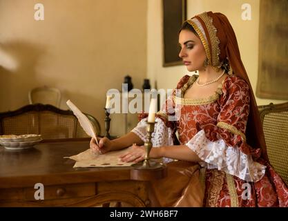 Frau im Renaissance-Kleid und französischer Kapuze, die einen Brief an ihren Tisch schreibt. Stockfoto