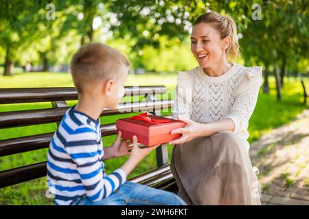 Glückliche Mutter sitzt mit ihrem Sohn auf einer Bank im Park. Der Junge schenkt seiner Mutter ein Geschenk. Stockfoto