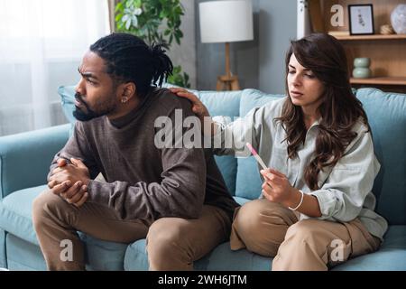 Unglückliche junge Frau, die auf dem Sofa sitzt und einen Schwangerschaftstest hat, ein afroamerikanischer Ehemann, der sie anschrie, drinnen. Millennial Familie hat Konflikt über un Stockfoto